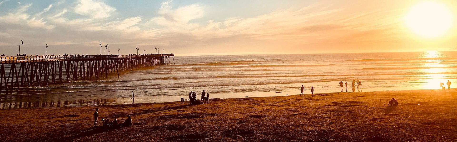 Sunset over a calm beach with a wooden pier stretching into the sea, silhouetted people strolling and playing on the shore.