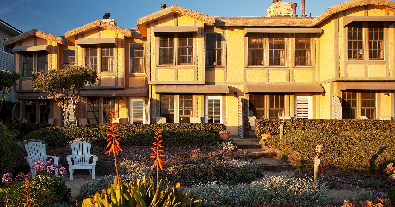 A row of beige townhouses with front gardens, sunlight glow, and colorful outdoor chairs in a tidy yard.