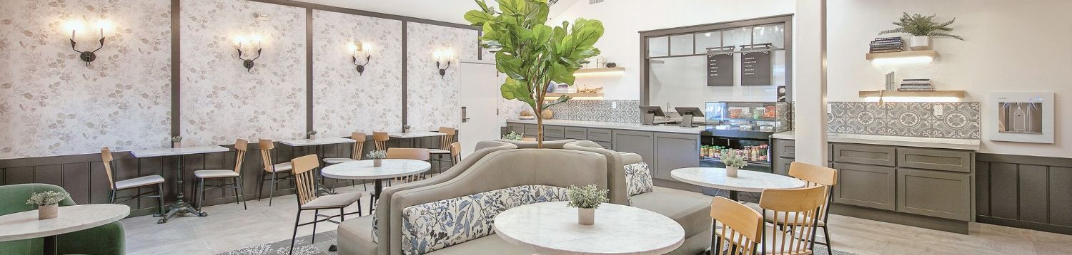 A modern cafe or lounge with round marble tables, wooden chairs, gray cushioned seating, a front counter, soft lighting, and a potted tree centerpiece.