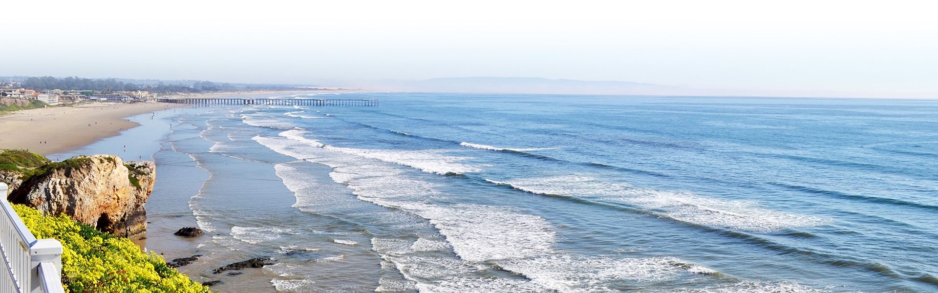 A sunny coastal scene with a long sandy beach, gentle waves, a rocky outcrop, and a distant pier extending into the ocean, under a clear sky.
