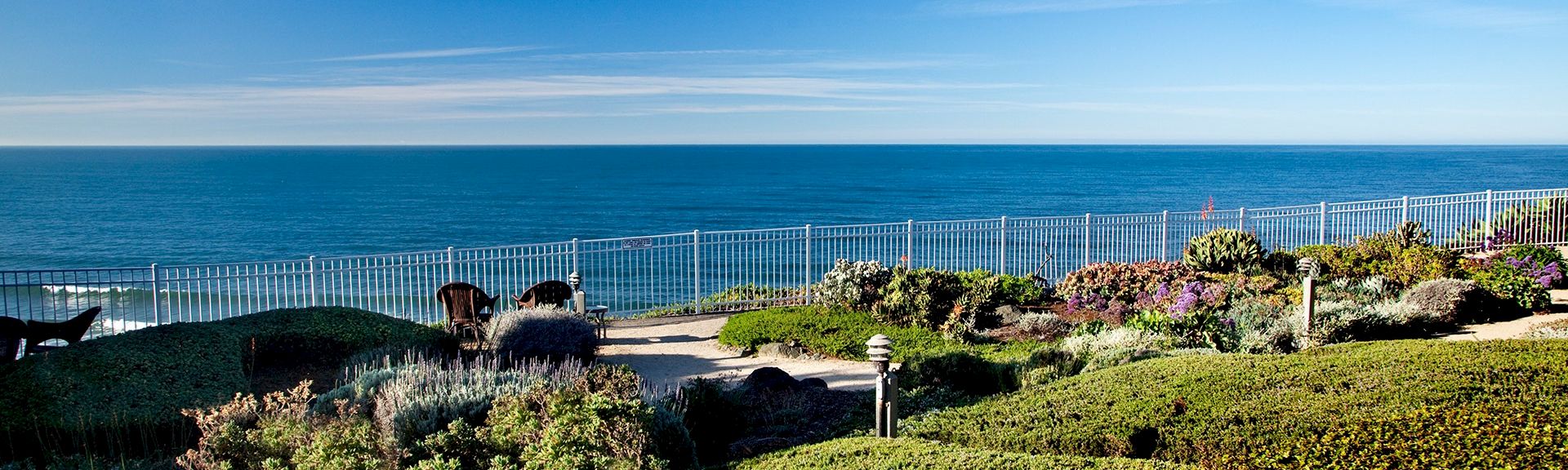 A bright coastal garden with neatly trimmed shrubs, a white fence, and a clear blue ocean stretching to the horizon behind.