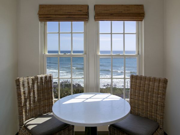 A cozy seaside breakfast nook with a round white table and two woven wicker chairs, overlooking the ocean through two large windows.