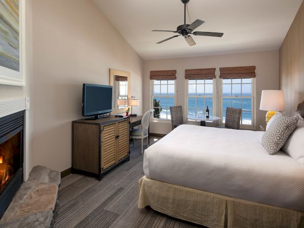 Cozy hotel room with a fireplace, king bed, desk, and a large window view of the ocean; warm lighting and neutral decor create a relaxing retreat.