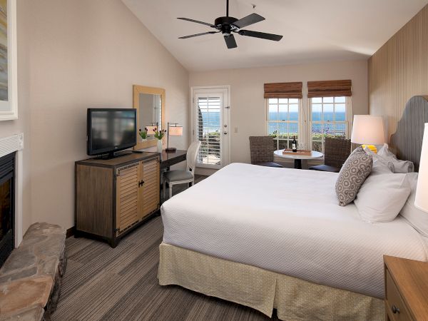 A hotel room with a king bed, fireplace, TV on a dresser, ceiling fan, two lamps, a seating area by the window, and a balcony view.