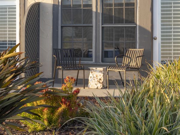 A small porch with two metal chairs and a floral-patterned chair, a windowed door, and surrounding garden plants.