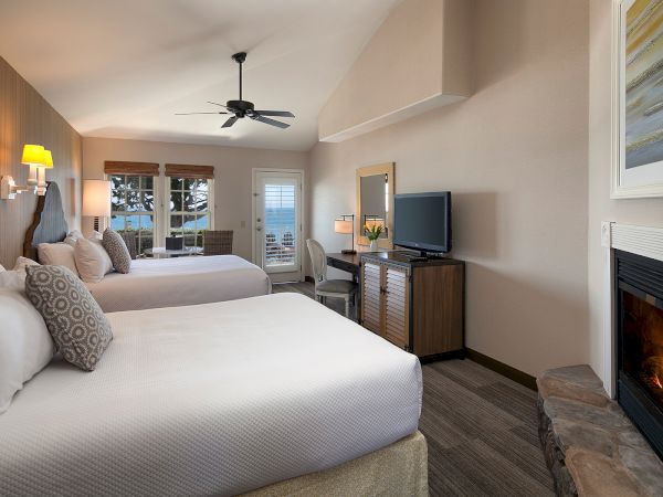 Two queen beds in a cozy hotel room with a fireplace, wall-mounted TV, desk, seating area, warm lighting, and a ceiling fan.