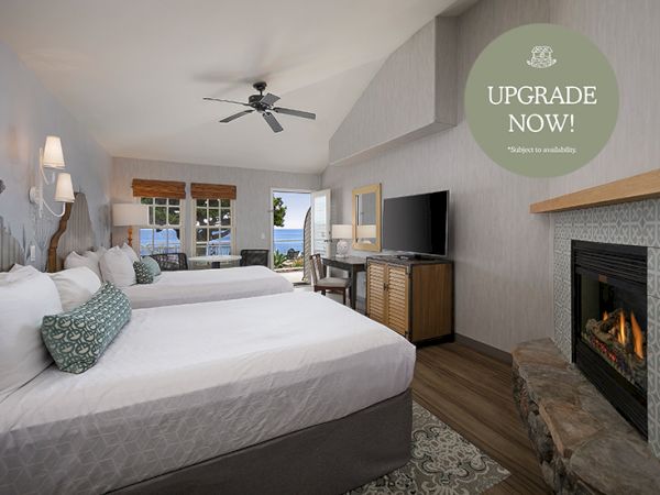 A bright hotel room with two plush beds, a fireplace, a flat-screen TV, and a balcony view; cozy seating and warm lighting, upgrade sticker.