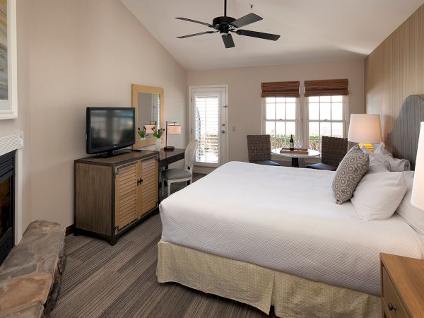 A cozy hotel room with a large bed, fireplace, TV on a dresser, a bench, and a seating area by windows; warm lighting and contemporary decor.