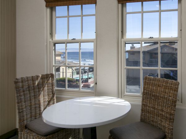 Cozy seaside cafe with a round white table and two wicker chairs by sunlit windows, blue sea visible beyond.