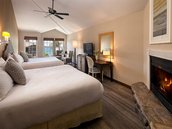 Two neatly made hotel beds, a desk area with chair, a fireplace, and a small sofa near a windowed door, all in a cozy, well-lit suite.