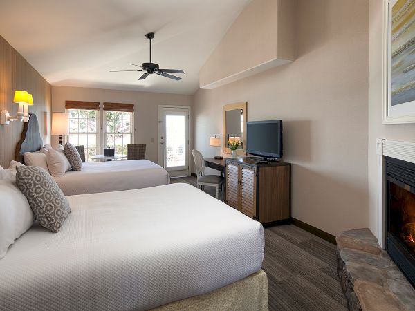 Two queen beds in a bright hotel room, with a fireplace, wall-mounted TV, desk area, ceiling fan, and a door to a balcony or outside.