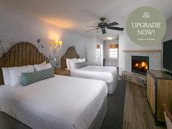 A hotel room with two queen beds, a ceiling fan, a fireplace, a TV stand, and a doorway, plus a round &ldquo;Upgrade Now!&rdquo; badge in the corner.