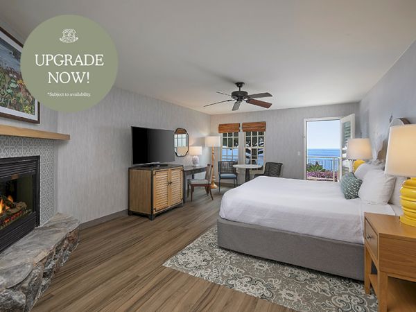 A bright hotel-style bedroom with a fireplace, a large bed, a dresser with a TV, a desk, and a balcony view, plus a ceiling fan and warm lighting.