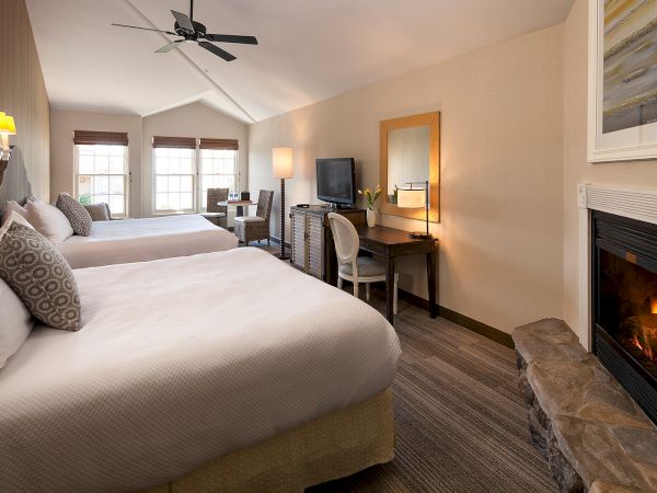 A cozy hotel room with two beds, a fireplace, desk and TV, warm lighting, and a large window seating area, all in soft neutral tones.