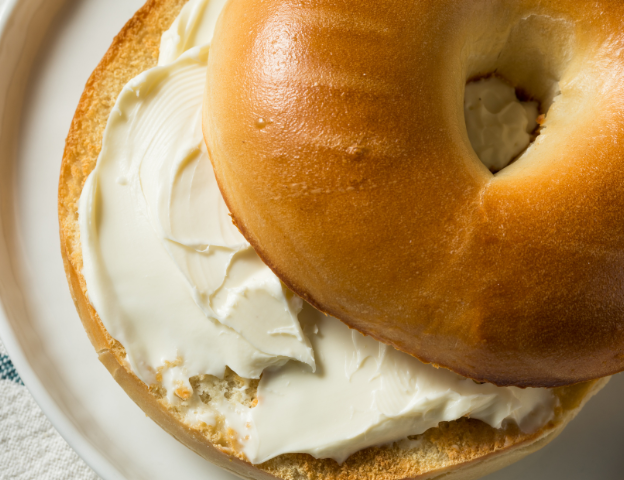 A bagel with cream cheese on a plate, photographed from above, showing a golden-brown toasted bagel half with generous white cream cheese.