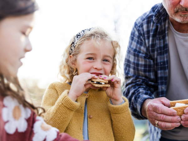 Three people share a sunny moment: a girl in a mustard sweater bites into a sandwich, while another child and an adult help nearby, outdoors.