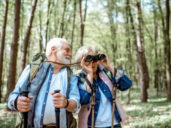 Two seniors hiking in a forest; one smiles with trekking poles while the other looks through binoculars, enjoying nature.