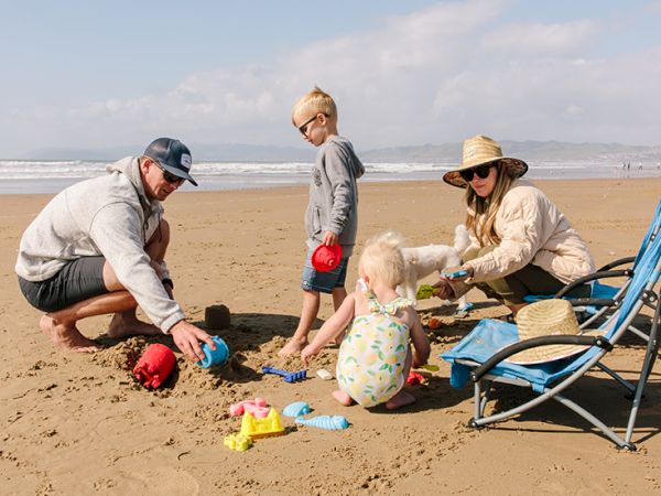 A family digs in the sandy beach with toys and a baby playing, while parents supervise near a beach chair by the waves.