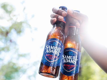 Two hands clink bottles of Samuel Adams beer outdoors, sunlight backlights the scene, celebrating with cold, amber craft brews.