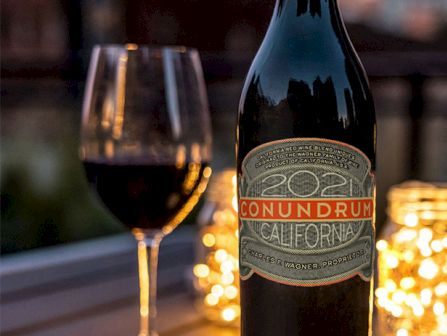 A bottle of wine labeled &ldquo;Conundrum&rdquo; with a glass of red wine and candles glowing softly in the background.