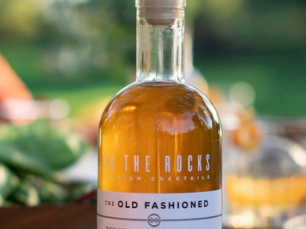 A bottle labeled &ldquo;The Rocks Old Fashioned&rdquo; on a blurred outdoor bar table with greenery and glasses in the background.