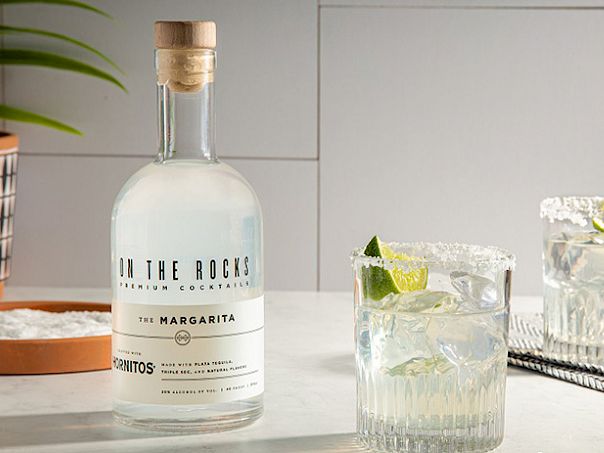 A bottle of vodka labeled &ldquo;On the Rocks&rdquo; beside two glass cocktails with ice and lime wedges, a plant pot, and a bowl of limes in a bright, modern kitchen.
