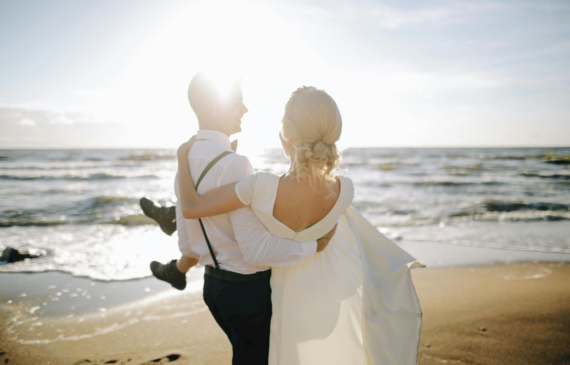 Two people on a sunny beach, a couple dressed in wedding attire walking along the shore with the man carrying the woman, waves in the background.