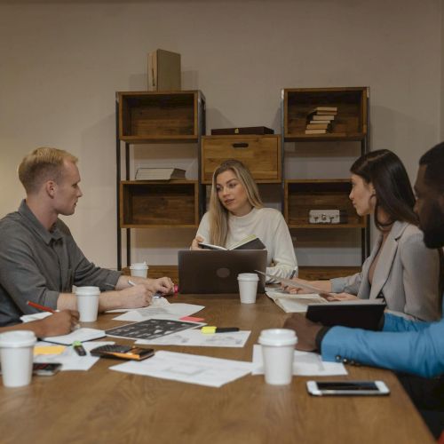 A diverse group sits around a table in a meeting room, discussing documents and laptops, with a woman presenting at the center.