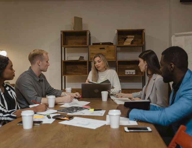 A diverse group sits around a table in a meeting room, discussing documents and laptops, with a woman presenting at the center.