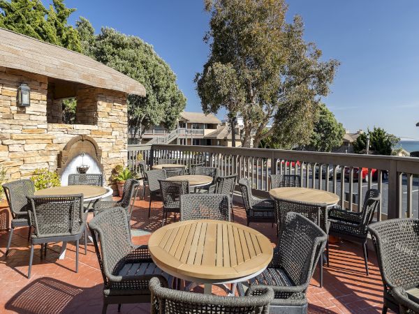 Outdoors cafe on a terrace with round wooden tables and wicker chairs, a stone fireplace, railings, trees, and buildings in the background.