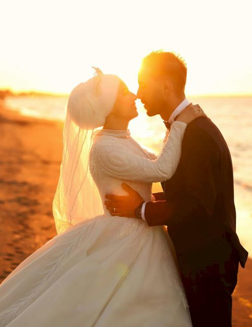A newlywed couple share a kiss on the beach at sunset, embracing in wedding attire with warm, golden light.
