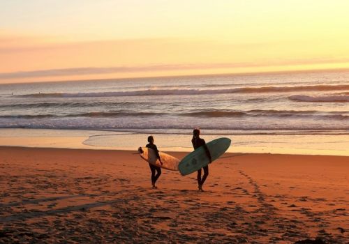 Two surfers walk along a sunset beach carrying boards toward the waves, footprints trailing behind them. (Done in 140 chars)