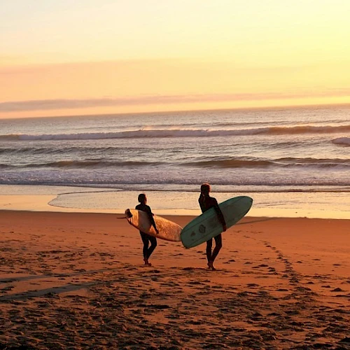 Two surfers walk along a sunset beach carrying boards toward the waves, footprints trailing behind them. (Done in 140 chars)