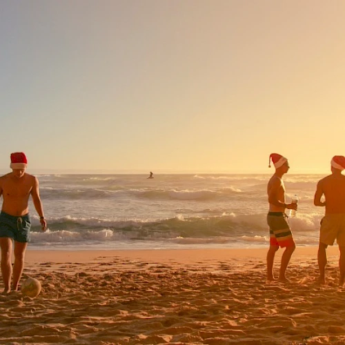 Three people on a sunny beach at sunset; two chat near the shore while a man walks the wet sand, gentle waves behind them.