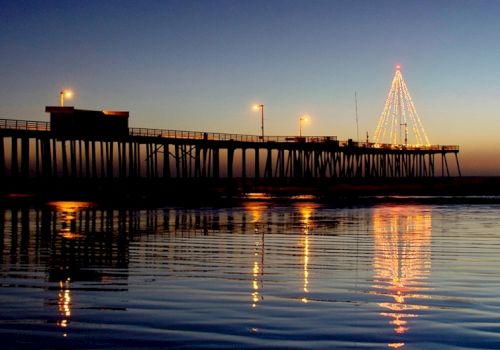 A wooden pier extends into calm water at dusk, lit lamps and a lit tall Christmas-tree-like structure shimmer on the surface.