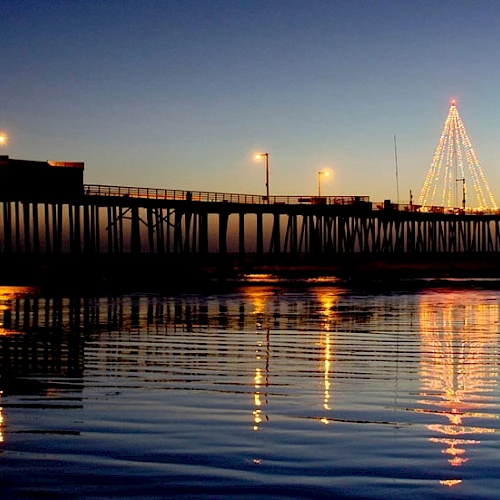 A wooden pier extends into calm water at dusk, lit lamps and a lit tall Christmas-tree-like structure shimmer on the surface.
