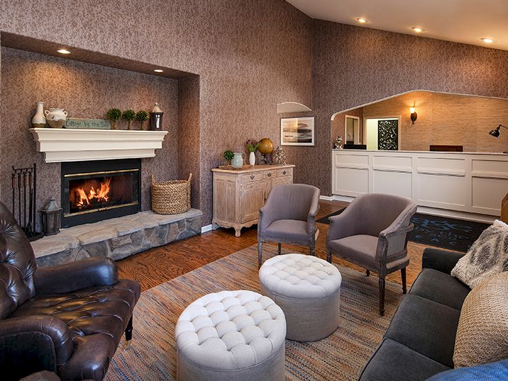 Cozy living room with a stone fireplace, warm wood floors, and comfy seating around two tufted ottomans, plus a bright kitchenette in the background.