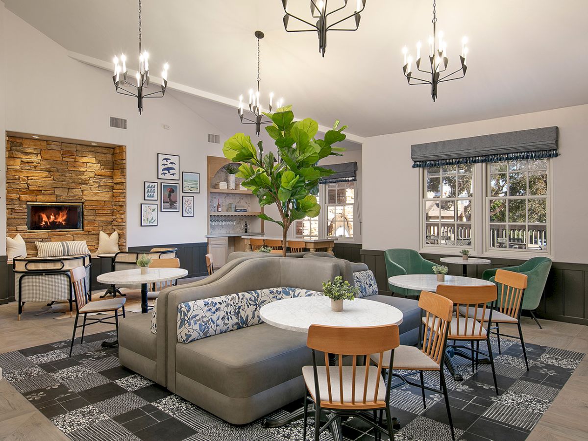 A cozy, modern lounge with a stone fireplace, greenery, and mixed seating around round tables. 140 characters, ending with a period.