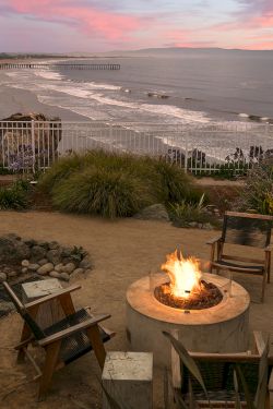 A seaside fire pit area with a circular fire pit and wooden chairs overlooking a beach at sunset, with a railing and plants nearby.