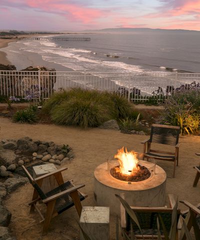 A seaside fire pit area with a circular fire pit and wooden chairs overlooking a beach at sunset, with a railing and plants nearby.