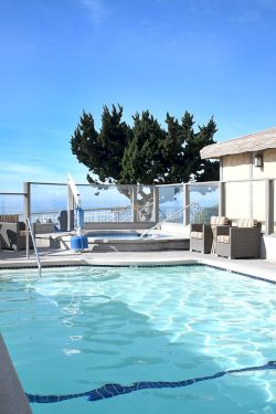 A rooftop hotel pool with clear blue water, lounge chairs, umbrellas, and a cityscape view, framed by a beige building and blue sky.