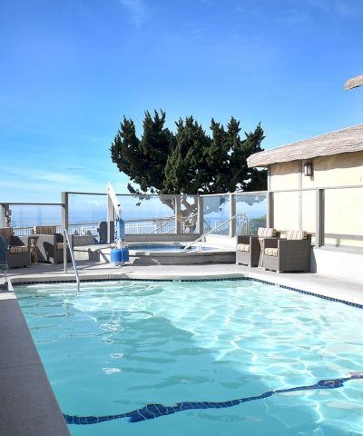 A rooftop hotel pool with clear blue water, lounge chairs, umbrellas, and a cityscape view, framed by a beige building and blue sky.