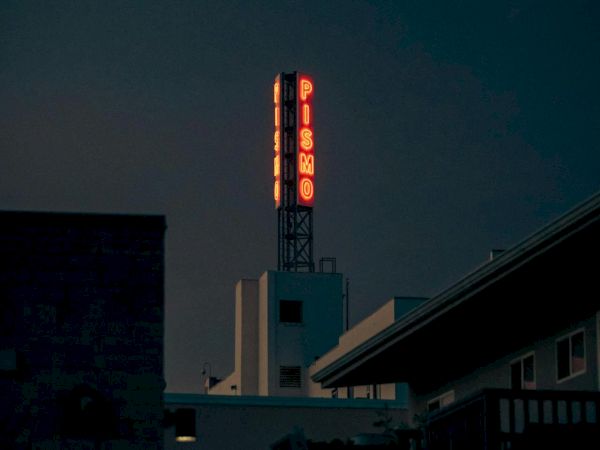 A dark city scene at dusk with a tall neon sign atop a building, glowing reddish-orange letters, perched above rooftops.