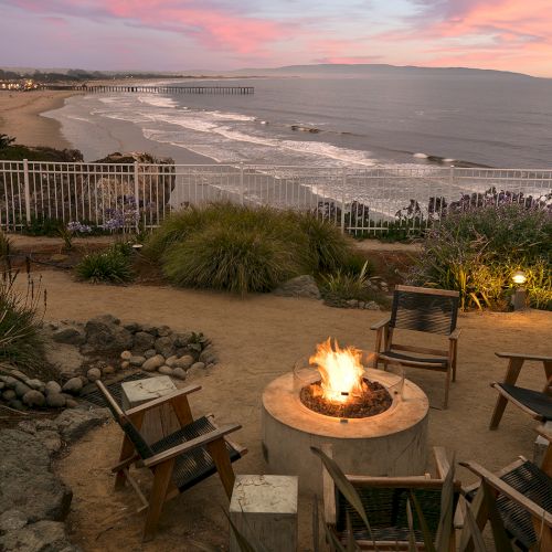 Sunset views over a terraced backyard with a stone patio, fire pit, and distant hills. A cozy, golden outdoor scene.