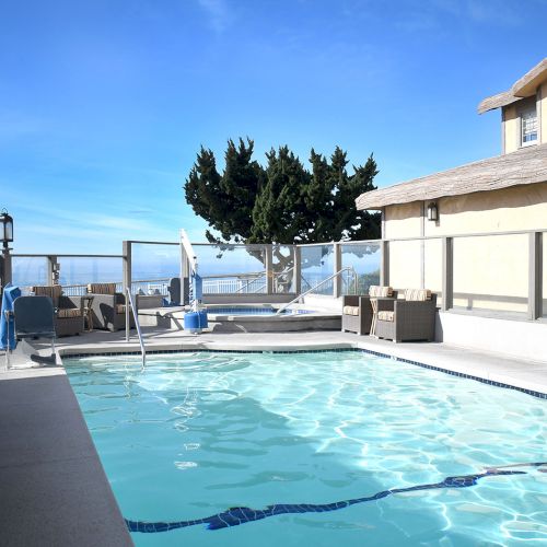 A sunny backyard with a sparkling pool, lounge chairs, and a modern two-story house overlooking the water, framed by palm trees and blue skies.