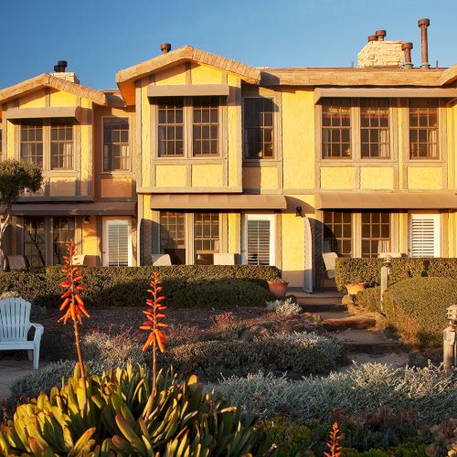 A beige hillside mansion with orange-tiled roof, balconies, lush greenery, and a sunny courtyard&mdash;evoking resort-like luxury and Mediterranean vibes.