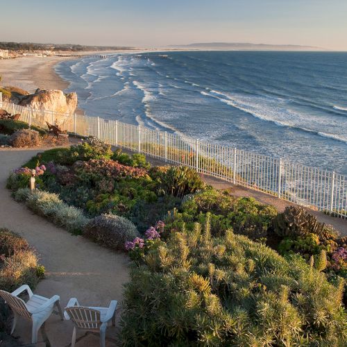 A rocky coastline with a lighthouse cliff path overlooking the ocean, waves crashing below, and a sunny sky with light clouds.