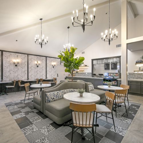 A modern, airy cafe or lounge with gray and green accents, comfy seating, patterned tile rugs, chandeliers, and a functional kitchenette area.