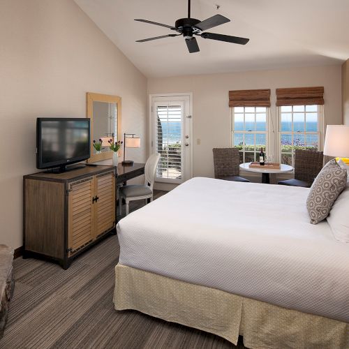 A cozy hotel bedroom with a large bed, soft lighting, a flat-screen TV, a small seating area, and a warm, neutral color palette.