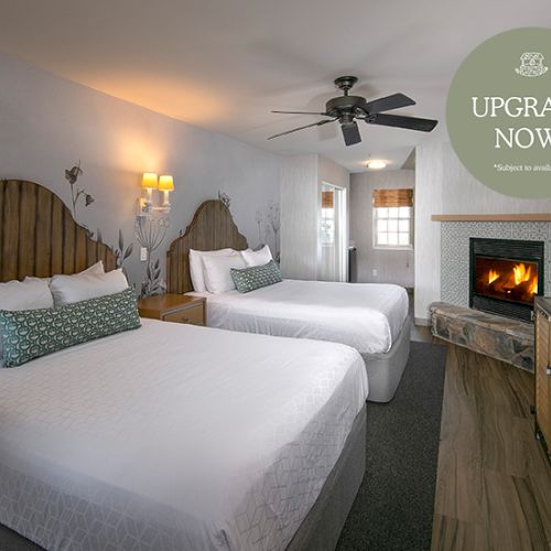 A cozy hotel room with two queen beds, soft lighting, a bedside lamp, and a small wooden desk; warm, inviting decor.